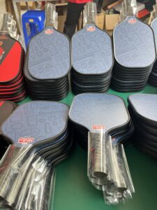 Pickleball paddle production workshop showing large batches of custom carbon fiber paddles in multiple colors, ready for assembly and packaging from a professional manufacturing factory