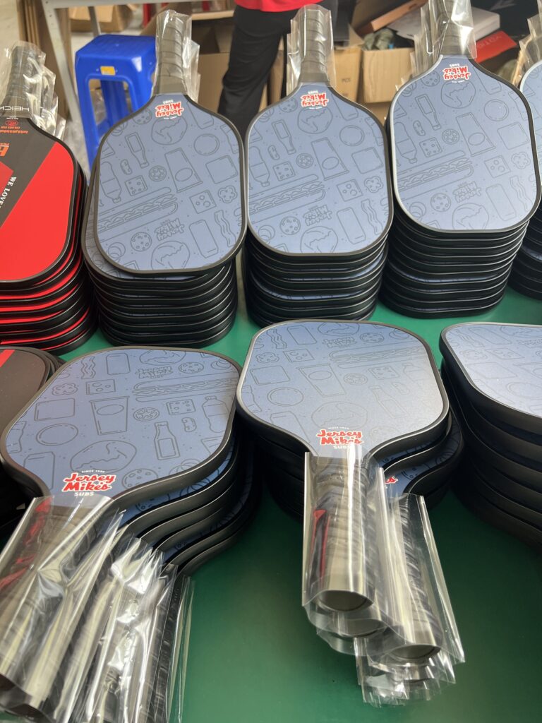 Pickleball paddle production workshop showing large batches of custom carbon fiber paddles in multiple colors, ready for assembly and packaging from a professional manufacturing factory